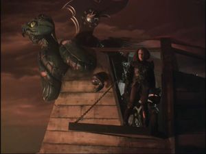 Under a sky full of red clouds, B'Elanna is standing on board a ship, looking out. The ship is old, made of wood, and its figurehead is a bronze griffin.