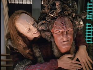 On an alien spaceship, Seksa, a white woman with Cardassian features,  is smiling and embracing a serious-looking Kazon man, Maje Culluh.
