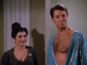 Troi is trying not very hard not to laugh at Riker, who is wearing a skimpy silk lamé outfit open to the waist, revealing one nipple and a generously hairy chest.