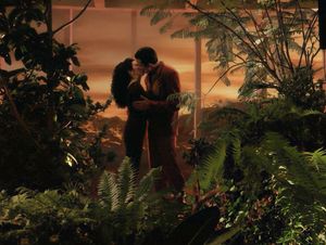 Deanna and a young man are standing in a garden in front of a window looking out onto a hostile alien landscape. They are kissing.