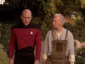 Captain Picard walks alongside an older man in gardening overalls, in a lush garden. The older man turns to him and says something.