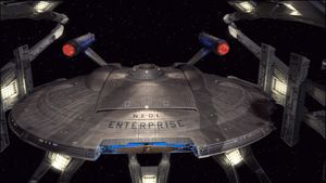The NX-01 Enterprise docked at an automated repair station, viewed from above and in front, with visible damage on one side of the saucer section and the ship's name and registry clearly visible.