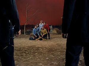 In the distance, the Enterprise Crew and Sylvia kneel over Chekov's body. They are framed by two enormous dark figures in the foreground.