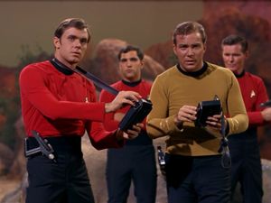 A cursed away team, consisting of Captain Kirk and three redshirts, stand on the surface of a TOS-era alien planet. One of them, a striking young man, is brandishing his tricorder.