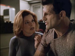 Captain Janeway is sitting next to Inspector Kashyk, a handsome man in a tight short-sleeved shirt. She is looking into his eyes and touching his upper arm. 