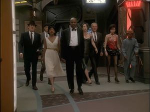 The crew of DS9, dressed in natty evening wear (apart from Nog, who's a janitor), walk along to promenade towards Quark's.