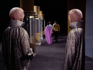 In the middle distance, Kirk, Spock, Bones and a beautiful but completely passive woman are walking past a row of perspex tubes into a dark void. Watching them, their backs to us, are two tall aliens with domed, hairless heads with visible buttock cleavage at the base of their skulls.