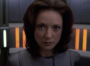 A young woman, Alice, in a slate grey jumpsuit is standing in a turbolift. She is looking at us with wide staring eyes.