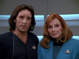 Dr Crusher is in the turbolift with a handsome man with a lot of latex on his forehead and 90s-era romance novel  cover hair. They are both looking at us with goofy expressions on their faces.