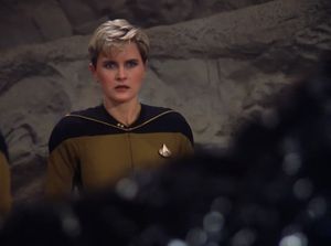 Tasha Yar is standing on the surface of an unconvincing alien planet. She looks concerned as a glistening black figure rises up in front of her.