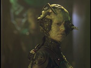Seven of Nine stands in Voyager's cargo bay. None of her implants have been removed yet: her skin is mottled, tubes come from her bare skull, and her left eye has been replaced by a spiky Borg sensor.