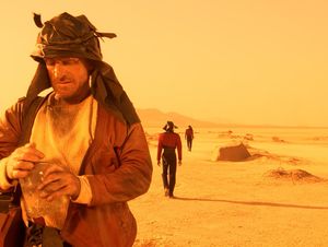 A grizzled middle-aged man screws the lid back onto a bottle of booze in the foreground, while Picard and Wesley walk away across a sun-scorched, barren landscape toward distant mountains.