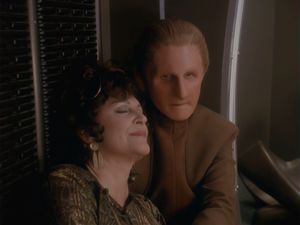 Odo is sitting against the wall of his quarters and Lwaxana is resting her head against his shoulder, her eyes closed in contentment. Odo's expression is unreadable.