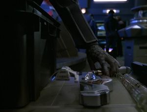 We're on the Defiant, in Engineering. Instrument panels are visible in the background. In the foreground, there's a table covered in futuristic instruments. An alien hand reaches out to grab one of the instruments, narrowly missing the tiny Danube-class runabout hovering above the table.