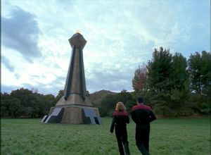 Janeway and Chakotay walk across a grassy field toward a tall stone war memorial, set among trees under a cloudy sky.