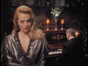Seven of Nine, wearing lipstick, a silver dress and her hair down, talks into a microphone. Behind her there is a pianist with his back to us.