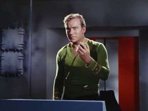 Captain Kirk stands in a corridor on a ruined space ship. He is talking into his communicator: he looks concerned.