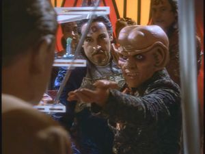 We're in Quark's Bar. Quark is holding his hand out to Odo, while a man with a moustache and some horrid forehead tattoos looks on. They are  standing around a perspex frame, which might just be a board game  of some kind.