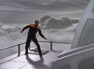 Tuvok, in his Starfleet uniform, stands in a wide-legged balancing pose on top of a maglev carriage, arms out from his sides, with a railing behind him and a bank of clouds stretching away behind.