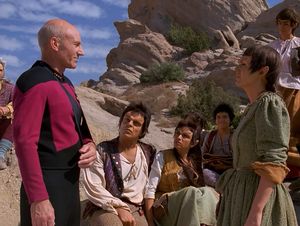 We're outside, on a sunny and rocky planet. Facing each other are  Captain Picard, his arm in a sling, and a woman roughly his age, wearing  a simple dress of rough fabric. Sitting between them and a bit behind them are a younger man and his daughter, also dressed in simple clothing. Picard and the woman are smiling at one another.