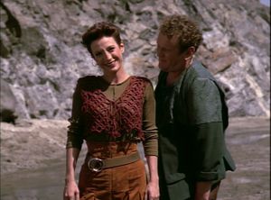 Kira stands in front of a rocky cliff face in a local quarry. She wears a cute outfit with a patterned red vest and a wide belt. O'Brien stands close behind her, leaning forward as if speaking quietly to her while she smiles toward someone off-screen.