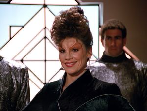 It's Ardra, a glamorous alien woman with dark hair piled on top of her head. She's wearing a dark futuristic dress and long dangling earrings. She's standing in front of a decorative window, flanked by guards. She smiles triumphantly.