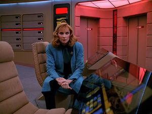 Beverly is sitting at ops on the bridge of the Enterprise. The red  alert lights are flashing. She is staring straight ahead, very intently.