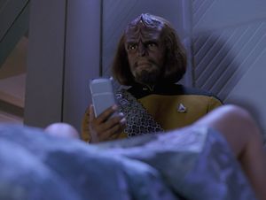Worf is standing in Ten Forward. He is holding out his tricorder, which he is using to monitor how dilated Keiko is as she is giving birth. Keiko's spread legs are visible in the foreground, covered decorously  with a shimmering space bedsheet.