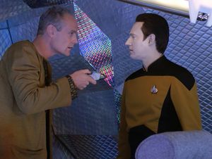 We are in a small shuttle, made to look futuristic with rainbow-coloured holographic wallpaer. A balding man with a big chin is pointing a phaser at Commander Data, who is completely unperturbed. 